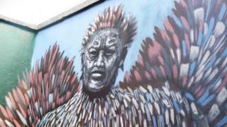 A vibrant mural fills the corner of an outdoor wall, showing a stylized human figure whose face is intentionally blurred or abstracted. From the figure’s head and shoulders, large feather‑like shapes burst outward in sweeping arcs of pink, blue, white, and brown, giving the impression of motion, energy, or transformation.