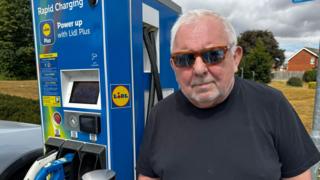 A man wearing sunglasses and a black T-shirt stands beside a blue Lidl Plus rapid electric vehicle charging unit at Lidl's Barton-upon-Humber store. Charging cables are connected to a vehicle parked alongside the charger. The unit displays instructions for using the charging service, while trees, housing and dark clouds are visible in the background.