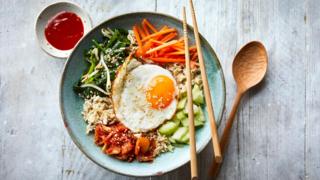 A blue bowl with cucumber, carrot, rice and kimchi, topped with a fried egg and pair of chopsticks