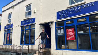 A man in shorts and a dark polo shirt stands at the entrance to a white building, with blue shop frontage advertising 'TAPS', 'SHOWERS' and 'GEO JONES & BROS' above three consecutive shop windows. There is a red 'Closing down sale' sign in the main window to the right.