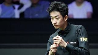 Wu Yize, wearing a black shirt and bowtie, chalks his cue with his left hand