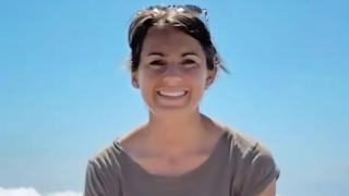 Family handout image of Lisa Ross - Close-up portrait of a person outdoors against a clear blue sky, wearing a grey short-sleeved top with sunglasses resting on the top of the head. The image is brightly lit by sunlight, with a small section of white cloud visible in the lower left corner.