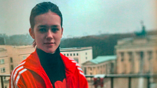 Brunette white teenage girl standing on a balcony. Background is blurred buildings. She is wearing a red sports jacket and black polo neck top.