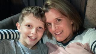 Jools (left) and Ellen (right) sit together in a chai, smiling for a selfie. Jools has short blonde hair and wears a grey top. Ellen has a blonde bob and wears a white, pink and navy striped jumper.