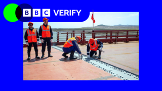 Four workmen wearing bright orange hats and reflective high-vis jackets stand at the centre of a new road bridge between Russia and North Korea. The centrepoint where the two ends of the bridge join is marked by a line of steel plates. Two of the workmen are crouched over the plates, examining them.