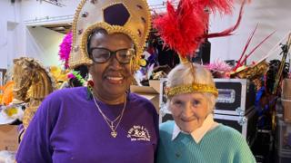 Two older women dressed in carnival head dresses smile together surrounded by carnival costumes and props
