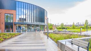 An wide paved road has a seat to one side and leads to a modern entrance of a railway station. It is a curved structure with long windows sitting above the doors and vegetation and grass on either side. A row of Victorian housing sits behind the station 