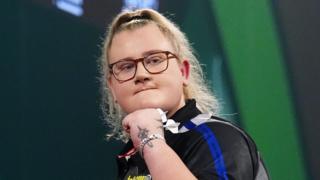 Darts player Beau Greaves holding two darts and looking into the distance wearing a black sports top
