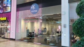 A glass-fronted polling station inside a shopping centre, marked with a circular “MK Votes” sign, with voting booths and a ladder visible inside.
