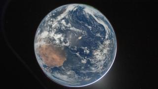 A picture of the Earth from space, which centres the planet against the dark background of space. It's a round blue planet. Clouds can be seen and a thin green aurora at the top.