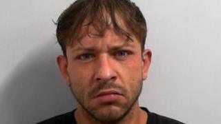 A mugshot of a man frowning directly at the camera in a police mugshot. He has a short dark beard and medium length brown hair. 