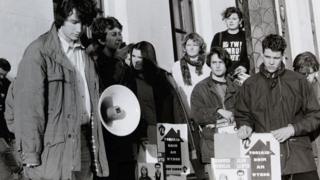 Mae'r llun du a gwyn yn dangos criw o bobl yn protestio o flaen yr hen Swyddfa Gymreig. Mae Rhun ap Iorwerth yn sefyll yno ac yn gwrando.