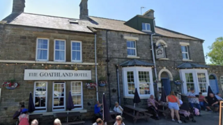The exterior of The Goathland Hotel, a stone-built two-storey structure built from grey stone, with a slate roof and several chimney stacks.
Above the entrance on the left-hand side is a large sign reading THE GOATHLAND HOTEL. The front of the building features large sash-style windows, stone detailing around the openings, and hanging flower baskets.
Multiple visitors are seated or standing in front of the building, with some gathered around the outdoor seating area.