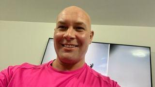 Kirk Marsden, who is bald, smiles posing for a photograph in a pink top. 