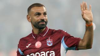 Mohamed Salah, visible from chest up wearing a claret Trabzonspor polo shirt with white and sky blue trim and a circular Turkish flag in the upper centre of the chest, raises his left hand to wave while showing a closed-mouth smile on his face