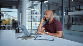 A picture of a man wearing a maroon polo shirt on the phone. He is at his desk with his laptop open.