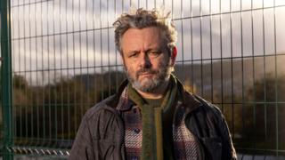 Michael Sheen looks at the camera. He is wearing a green scarf and a plaid red shirt and a coat over the top. Behind him is a waste site which is fenced off.