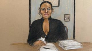 Court sketch of Gabriella Carrington who has pen on a piece of paper next to s stack of papers. She is in front of a blue prison door and is in black