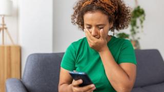 A woman in a green t-shirt holding her hand over her mouth in horror as she looks at her phone while sitting on a grey sofa.