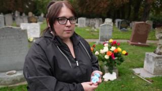 Jasmine Beverley wearing a black coat and black glasses holding her premature baby's small blue teddy bear. She is stood in front of her son's grave which is full of coloured roses.