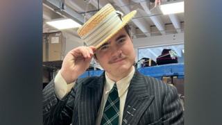 A person in a dark pinstripe suit, cream shirt, and green plaid tie stands indoors holding a straw boater hat. Behind them are shelves filled with hats and boxes in a workshop‑like space with exposed beams and fluorescent lighting.