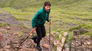 Trish Patterson, a woman with short hair walking with two walking sticks on a rocky terrain. She is wearing black trousers and a green coat and appears out of breath as she looks to the camera.