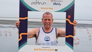 A runner in a white vest is holding up a cardboard photo frame that shows he is in Walvis Bay in Namibia. He is standing in front of a tidal lagoon and there are dozens of pink flamingos on the sand behind him