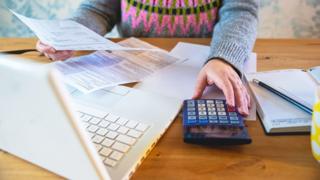 A woman looking at paperwork and typing on a calculator next to a laptop