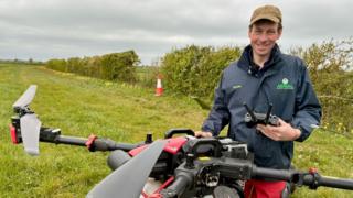 Frazer Bloor, wearing a beige baseball cap and navy jacket, stands behind a large red and black drone. In his hand is a black remote control. He is standing in a field.