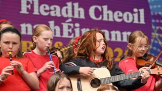 A group of young girls play instruments on a stage including a guitar, a fiddle, and tin whistles. They are standing on a stage in front of an Irish language sign which says Fleadh Cheoil na hÉireann.