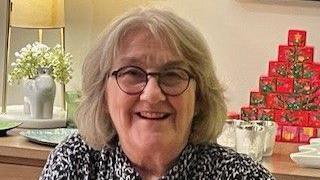 A headshot of a woman with short blonde, bobbed hair and glasses with black frames. There is a lamp behind her on a wooden sideboard as well as a red Christmas tree decoration and three silver candles. She is smiling and looking into the camera. 