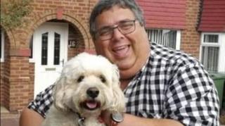 A man smiles at the camera, holding a fluffy white dog. He had dark grea hair and glasses, and is wearing a black and white checked shirt. Behind him is a brick house with the nuber 16 next to the front door.