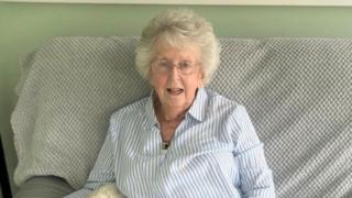 An elderly lady sitting on the sofa she has white hair and has a pair of round glasses on, she is wearing a blue and white stripped shirt and has a gold necklace on.