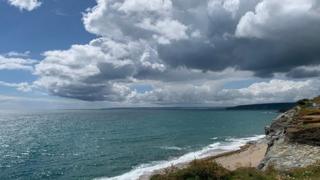 Clouds over a turquoise sea with cliffs and the shore in the foreground in Cornwall.