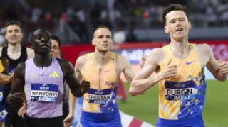 Max Burgin of Great Britain crosses the finish line to win the 800m race during the Diamond League athletics meeting in Rabat. Burgin, on the right-hand side of the image, is part of a group of athletes all seen from waist up running towards the line. He is dressed in blue shorts and a yellow and blue running vest with a bib with his name on the front, and has his hands high as he pumps his arms. On the left-hand side of the image, slightly behind, is Kenya's 800m Olympic champion Emmanuel Wanyonyi in a lavender running vest and name bib
