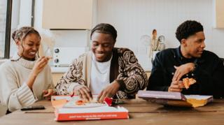 Three students sit in a kitchen eating pizza
