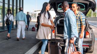 Stock photo shows a group of people removing luggage from the boot of a car as it pulls up to an airport with other passengers walking past.