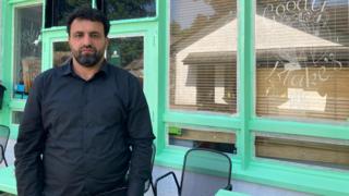 Shwan Khder is wearing a black shirt. He has a beard and looks unhappy. Behind him there is a building with large windows and light green window frames.