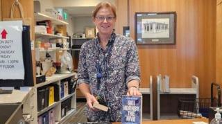 Joanne - a woman with straight pulled back hair, glasses and a floral print short is standing behind a library service desk holding a blue book, titled Gill Hornby The Hive