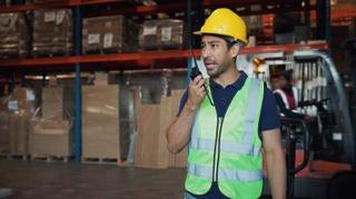 A picture of a man wearing a high-vis jacket and a yellow helmet speaking through a walkie talkie in a stock room.