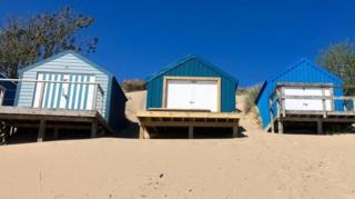 Three blue beach huts sitting high on a sandy beach and beneath a blue, cloudless sky