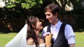 A young couple outdoors wearing wedding clothes. They are looking into eachothers' eyes and smiling. The bride is wearing a veil and holding a paper cone of flowers.