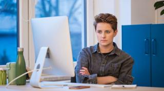 A young woman with short hair wearing a grey denim shirt working in an office, sitting with arms crossed at the desk in front on computer.
