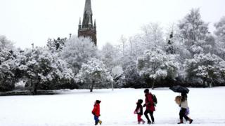 Snowy scene with a park area and a church in the background. Trees covered in snow and a group of people walking in the snow.