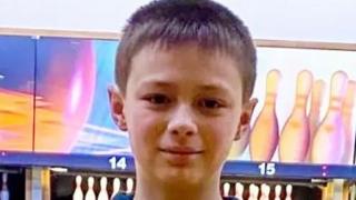 A close-up photo of a boy looking straight at the camera. He has short brown hair and he is wearing a blue hoodie. He is in a ten-pin bowling alley, with lanes and sets of pins visible behind him.