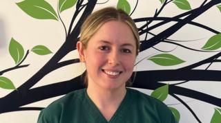 Lucy is wearing green NHS scrubs, with the NHS logo embroidered on the chest. She has blonde hair tied back and is smiling at the camera. She is stook against a white wall with a tree and leaves painted on it.