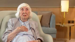 Betty Brown wearing a blue dress, grey jacket and pearls sits in a green armchair. In the background is a lamp on a table.
