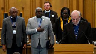 A clergyman at a lectern with some  black clergy standing behind him