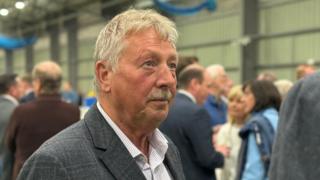 Sammy Wilson, a man with short, grey hair and a moustache, looks away from the camera towards a reporter during a TV interview. He is standing in an event centre during an election count. He is wearing a white striped shirt and a grey checked suit jacket.