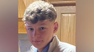 The image is a cropped photo of Connor Magee Campbell. He has short blonde hair and is wearing a blue shirt with a grey jumper.